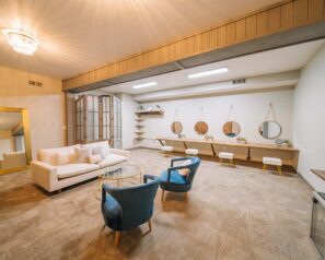 A large room outfitted with elegant modern furniture. There is a seating area, a large floor length mirror and a long dressing table on the far wall with spaces for 4 people - each with a short stool and large round mirror on the facing wall.